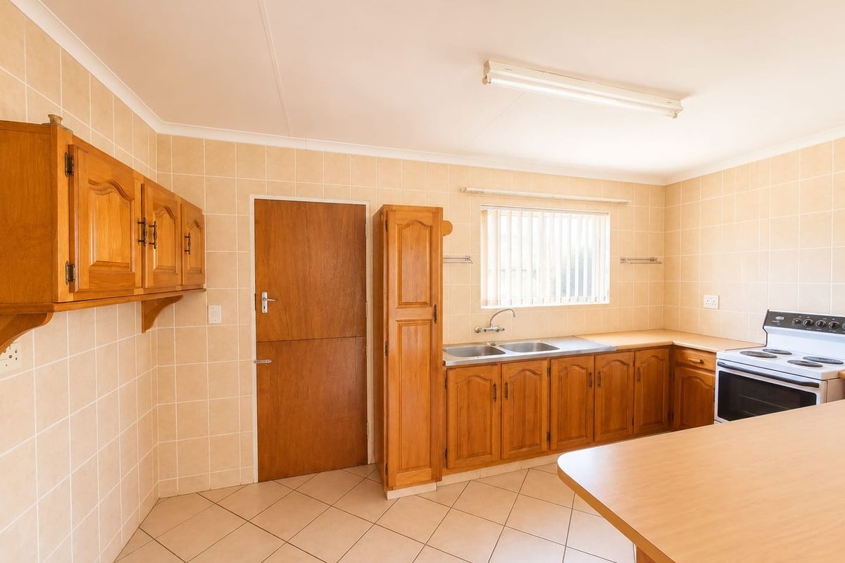 A functional kitchen featuring wooden cabinetry, laminate countertops, a double stainless steel sink, and a freestanding electric stove with neutral wall and floor tiling.