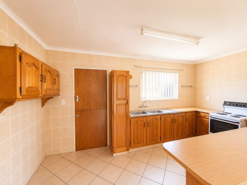 A functional kitchen featuring wooden cabinetry, laminate countertops, a double stainless steel sink, and a freestanding electric stove with neutral wall and floor tiling.