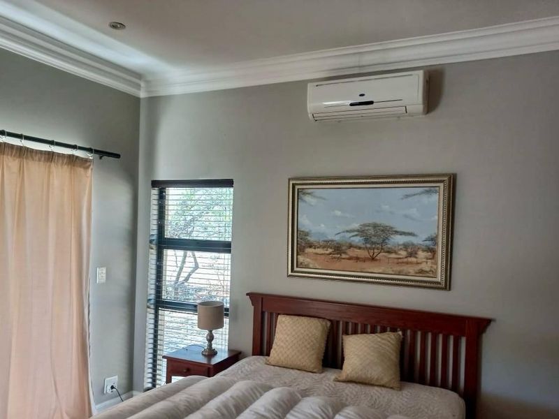 A well-maintained bedroom featuring neutral wall tones, crown molding, a wall-mounted air conditioning unit, and large windows with fitted blinds.