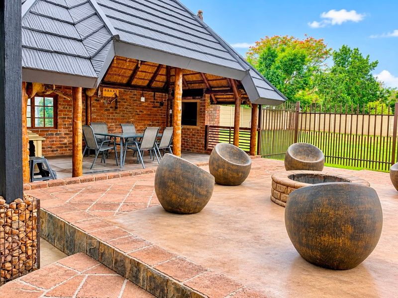 A spacious outdoor entertainment area featuring a covered thatched patio with a built-in braai, adjacent to an open stone-paved terrace with a fire pit.