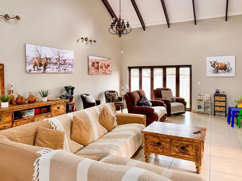 Spacious living room featuring high vaulted ceilings with exposed timber beams, tiled flooring, and large windows fitted with venetian blinds.