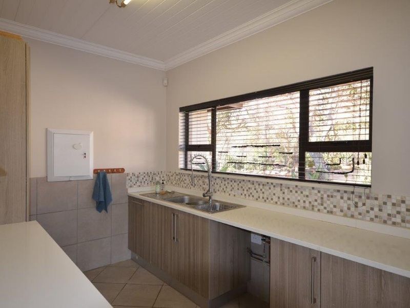 A clean and functional scullery featuring laminate cabinetry, a double basin sink, mosaic tile backsplash, and tiled flooring with natural light.