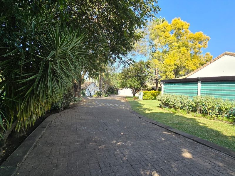 A well-maintained paved driveway leading towards the property, surrounded by established trees, lush garden beds, and privacy screening.