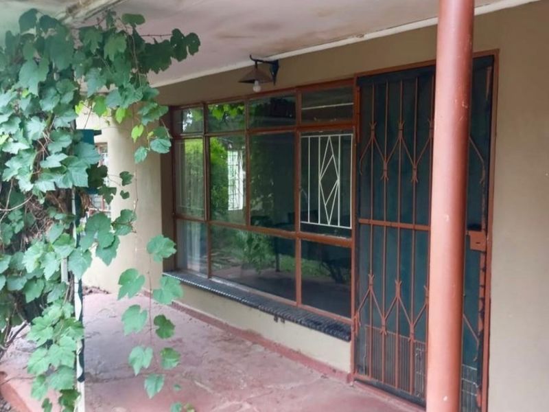 A covered patio area featuring large glass windows, a metal security gate, and a support pillar, showing signs of wear and needing maintenance.