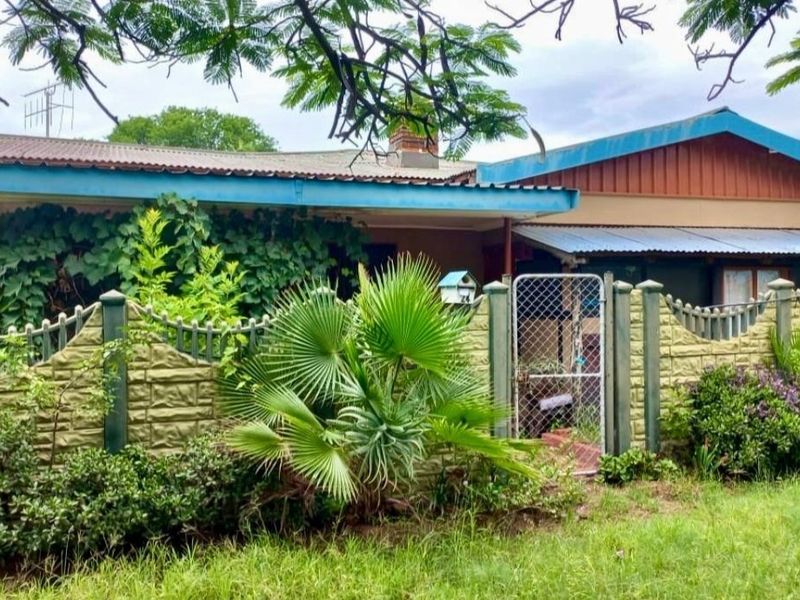 Front view of a single-story house featuring a precast concrete boundary wall, metal gate, and corrugated metal roofing with established garden surroundings.