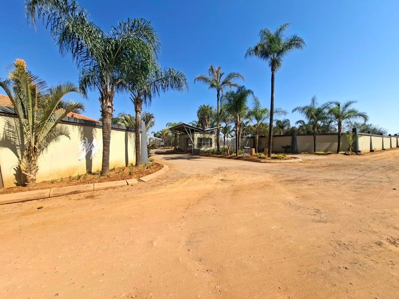 Entrance view of a residential estate featuring a security gatehouse, perimeter walling, and established palm tree landscaping.