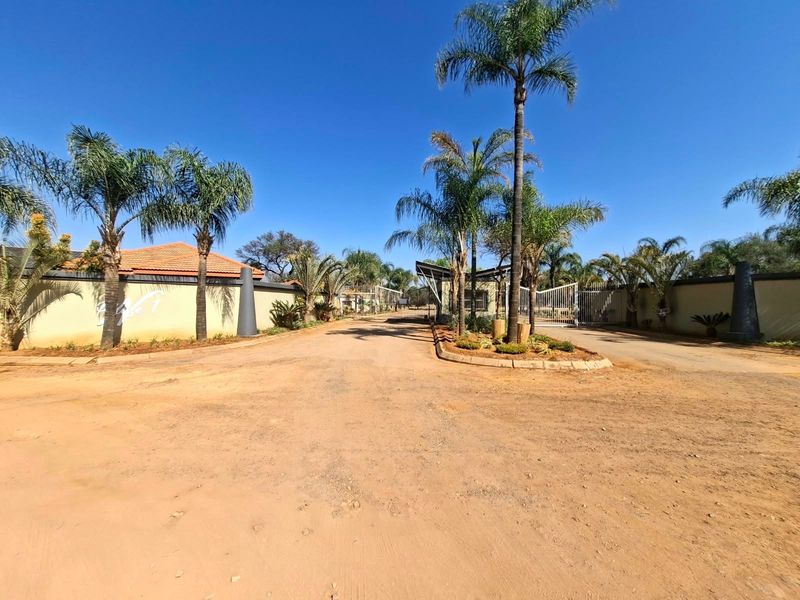 Entrance view of a residential estate featuring a secure gated access point, perimeter walling, and established palm tree landscaping.