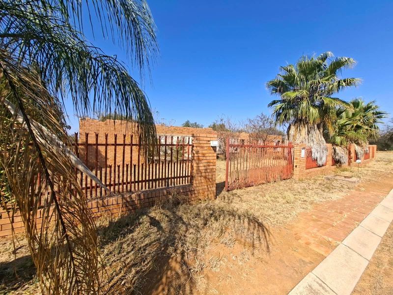 Street view of a residential property featuring a brick boundary wall, metal security fencing, and overgrown landscaping.