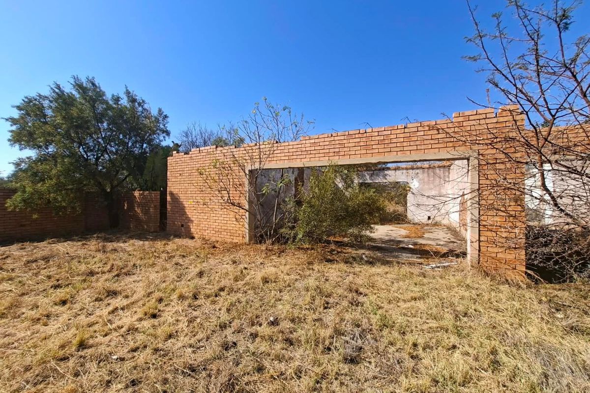View of an incomplete brick structure on a vacant residential plot, showing exposed masonry and overgrown grounds.