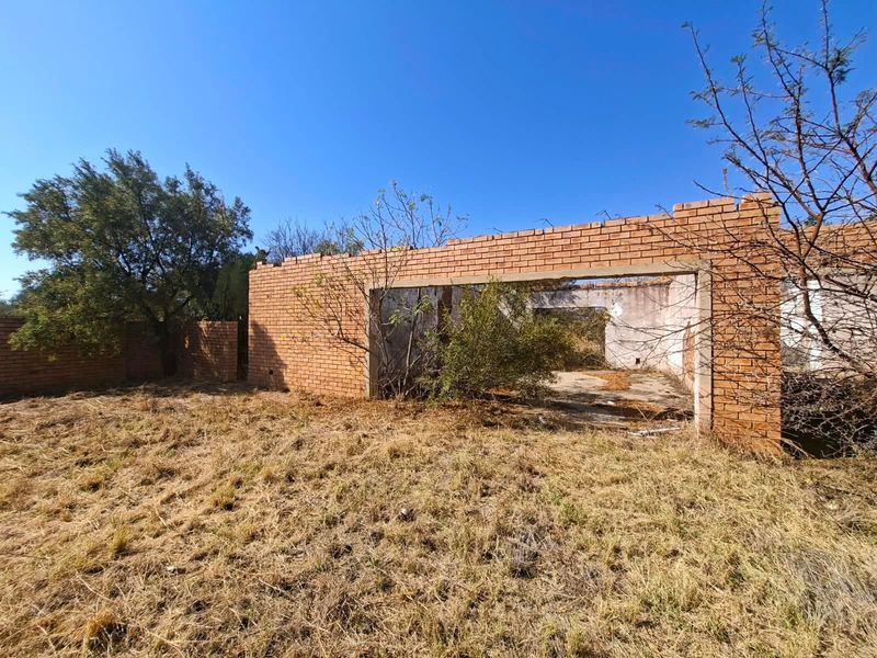 View of an incomplete brick structure on a vacant residential plot, showing exposed masonry and overgrown grounds.