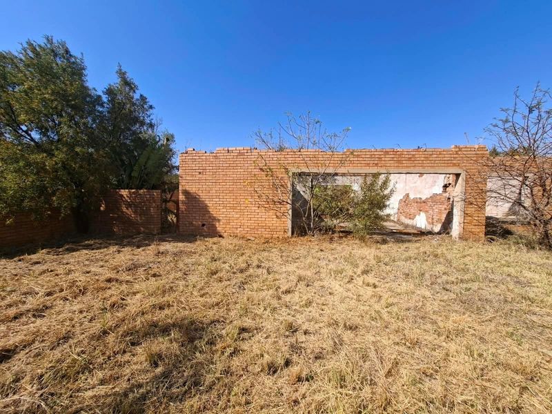 A view of a dilapidated brick structure on a vacant residential lot, showing significant structural deterioration and overgrown vegetation.