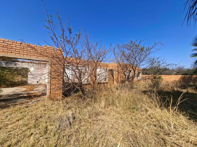 A derelict residential structure featuring exposed face brick walls and a concrete foundation, situated on an overgrown plot of land.