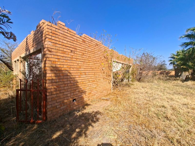 An exterior view of an incomplete brick structure on a vacant residential plot, showing exposed masonry and overgrown grounds.