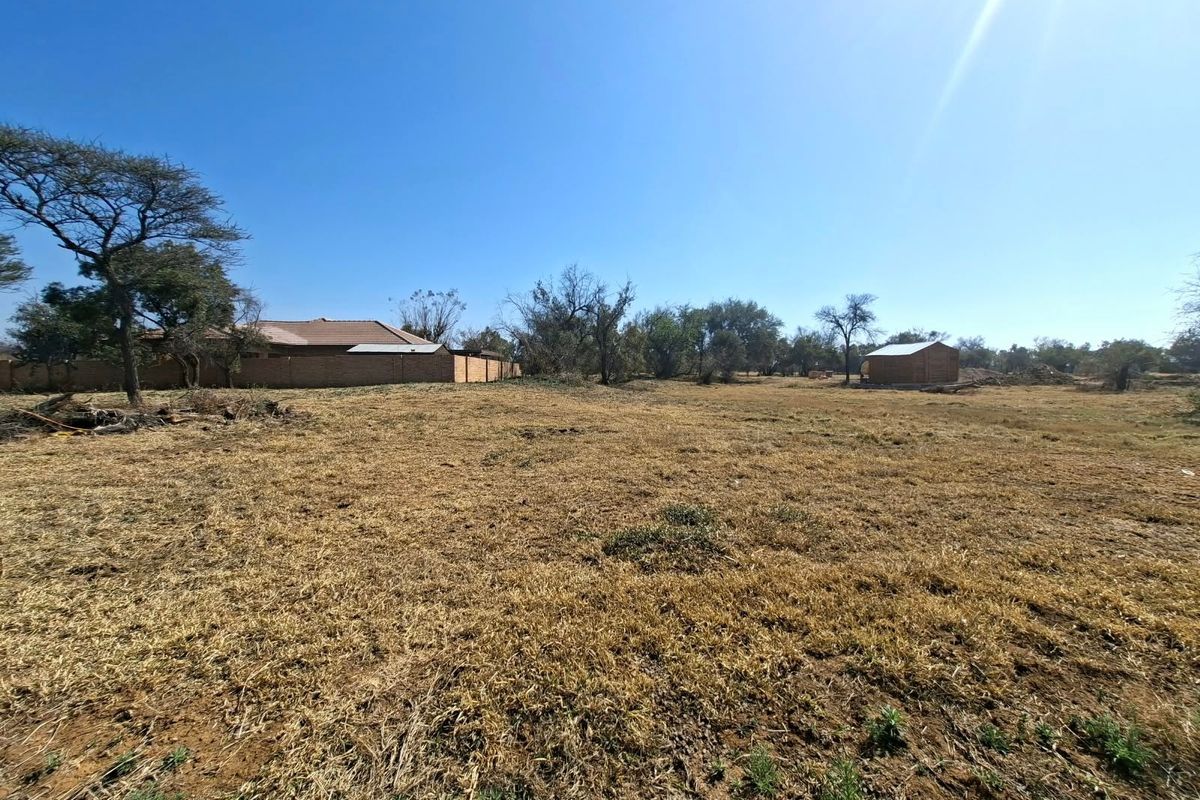 A wide-angle view of a vacant residential plot featuring dry grass, mature trees, a boundary wall, and a small outbuilding structure.
