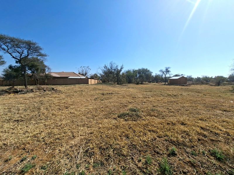 A wide-angle view of a vacant residential plot featuring dry grass, mature trees, a boundary wall, and a small outbuilding structure.