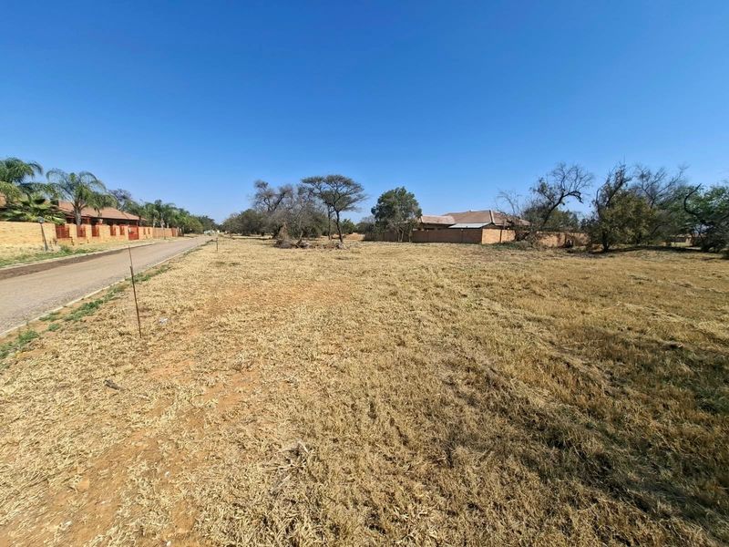 A vacant residential plot of land situated along a paved road, featuring natural grass cover and surrounding boundary walls.
