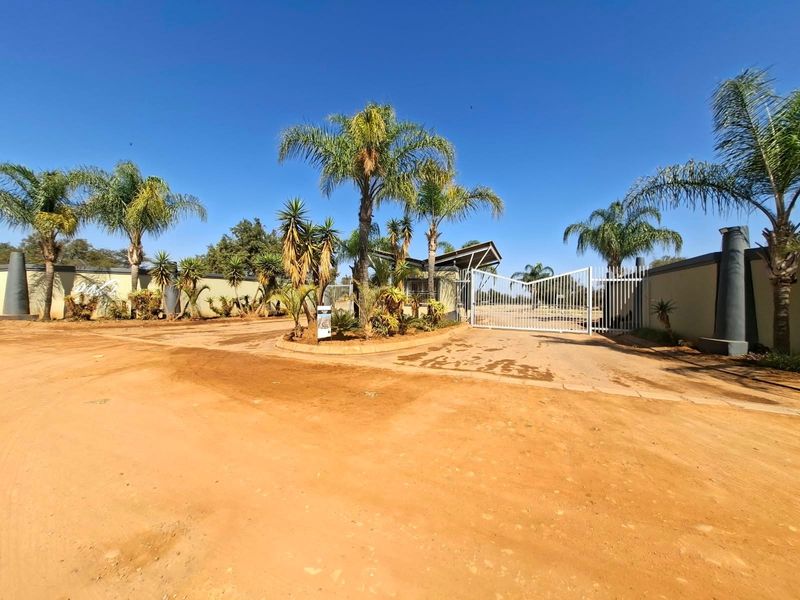 Front entrance view of a residential estate featuring a secure gated access point, perimeter walling, and established palm trees.