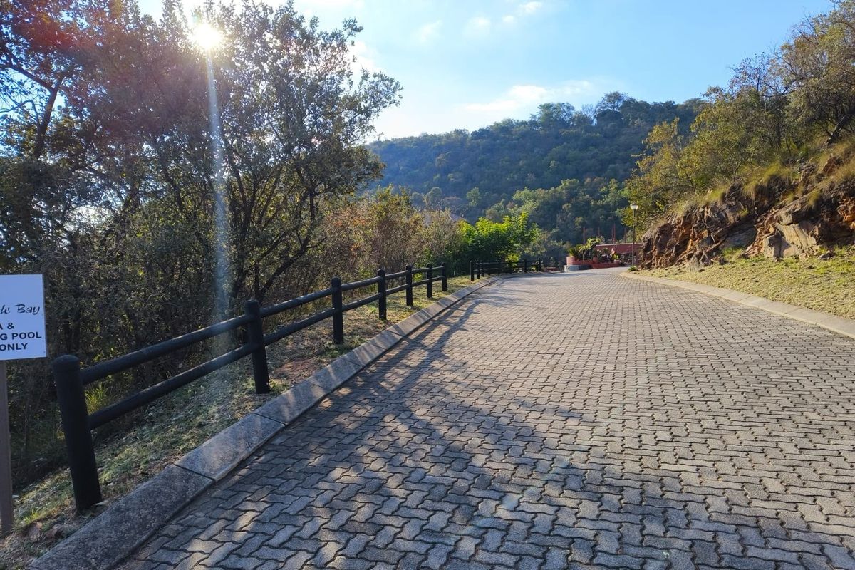 A paved driveway leading through a landscaped residential area, featuring timber post and rail fencing and natural hillside surroundings.