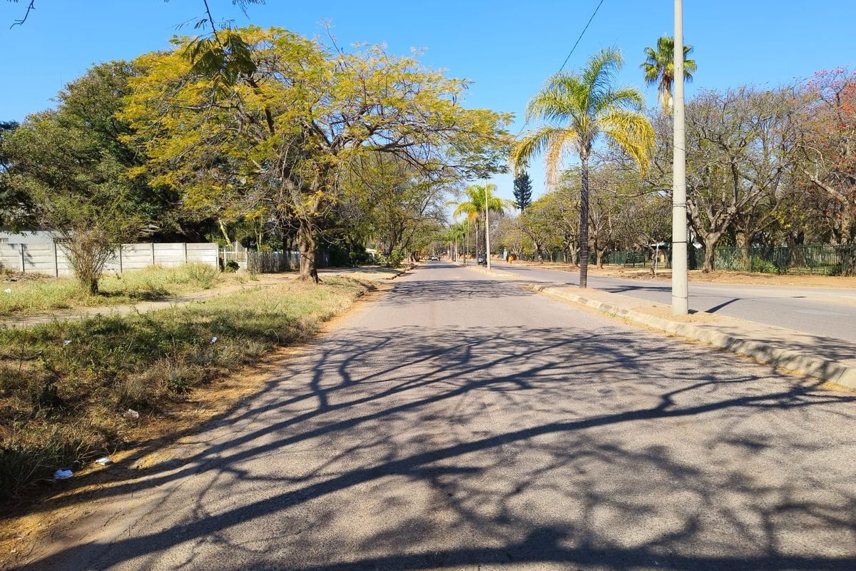 A view of the residential street frontage featuring mature trees, asphalt road, and established boundary walling in a suburban neighborhood setting.