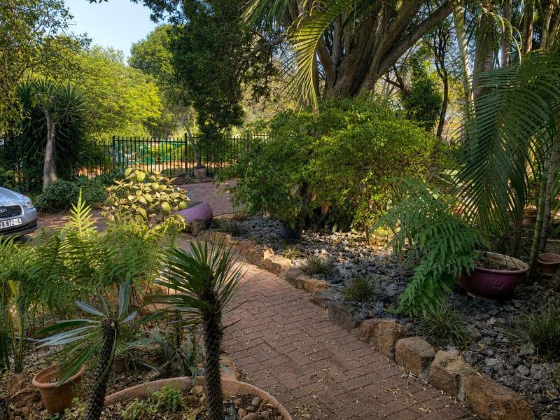 A lush, established garden featuring a brick-paved pathway, stone edging, and mature trees enclosed by a metal perimeter fence.