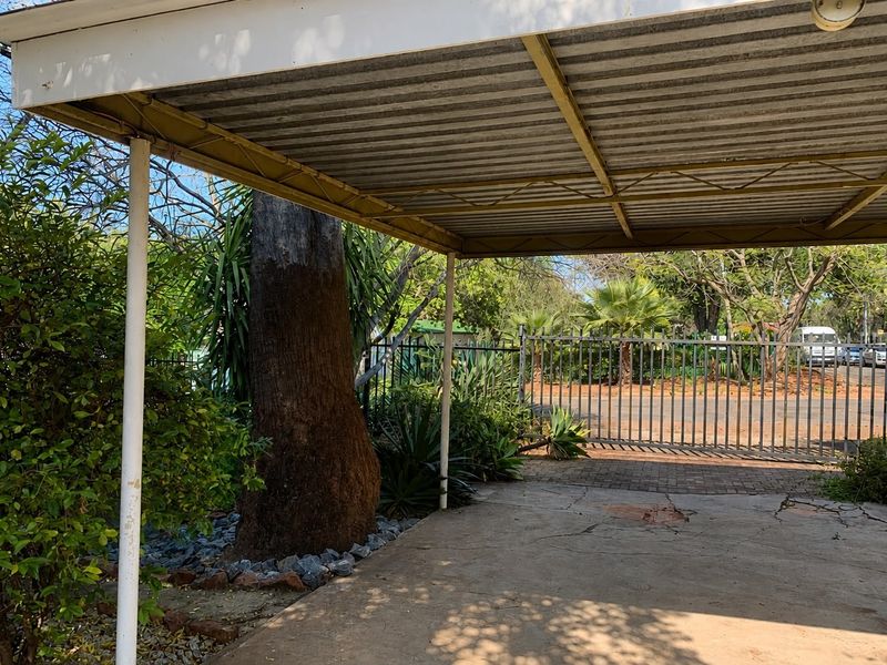 An open-sided carport structure featuring a corrugated metal roof, steel supports, and paved flooring, situated within a garden setting with perimeter fencing.