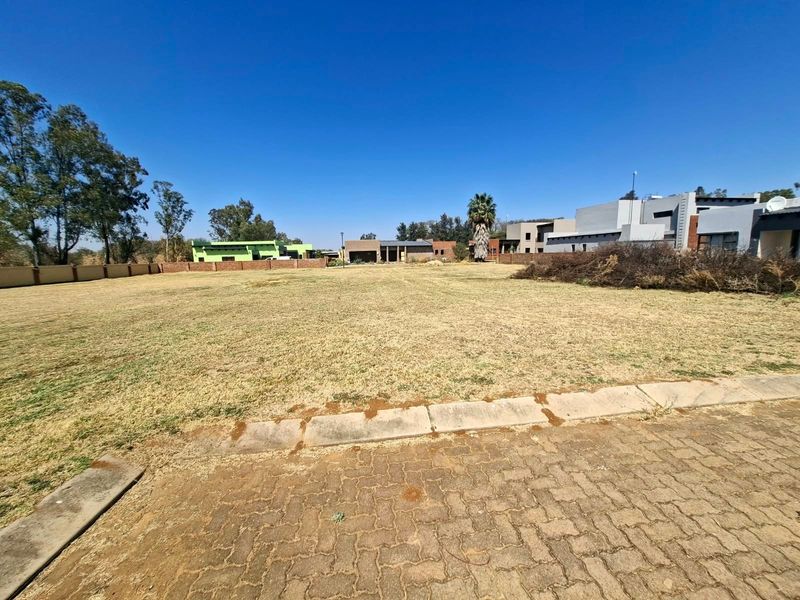 A vacant residential plot featuring a level grassed area, paved driveway access, and established boundary walls in a developed estate setting.