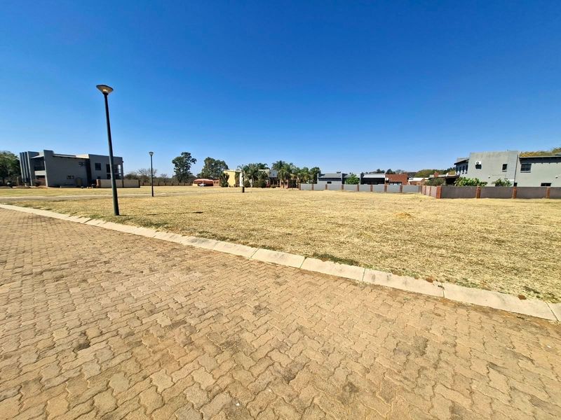 A vacant residential plot featuring paved street access, concrete curbing, and established street lighting in a developed estate setting.