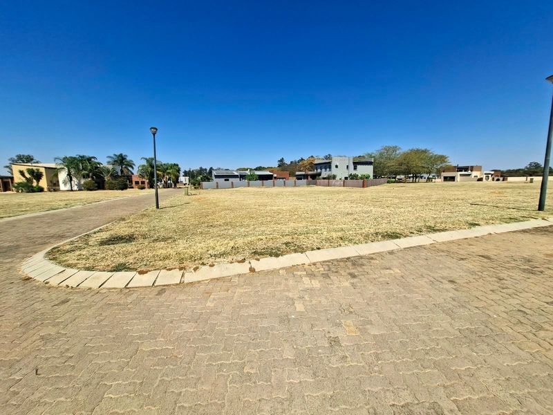 A vacant residential plot featuring established paving, kerbing, and street lighting, situated within a developed neighborhood setting.