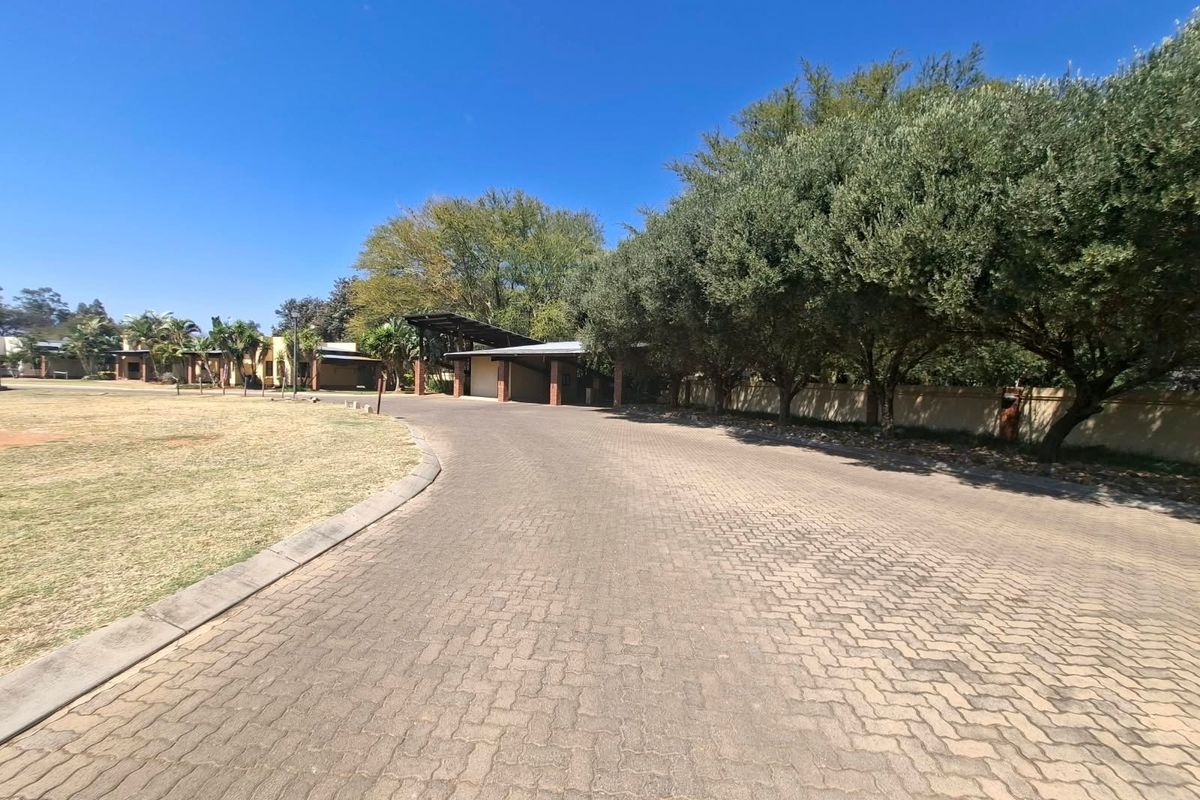 A wide paved driveway leading towards a covered carport structure, surrounded by mature trees and a perimeter wall on a residential property.