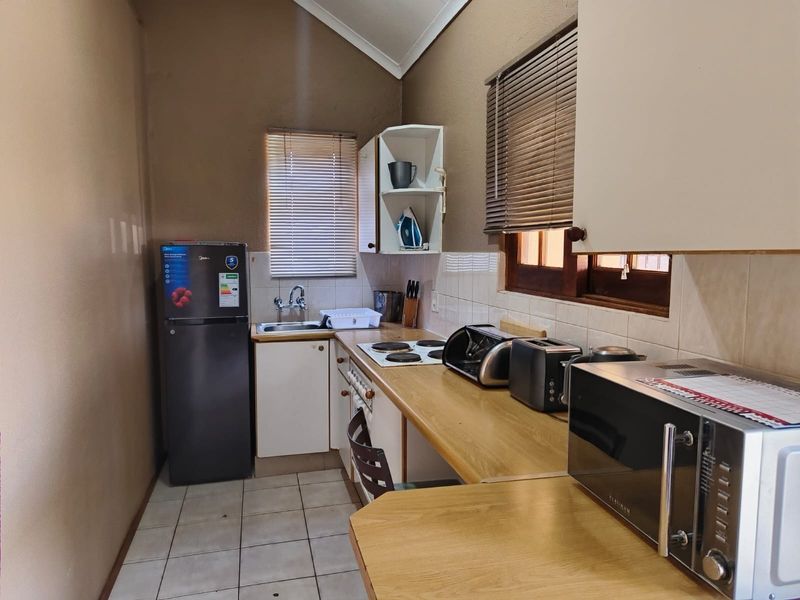 A compact kitchen featuring laminate countertops, built-in cabinetry, an electric hob, and a stainless steel sink with tiled backsplash and venetian blinds.