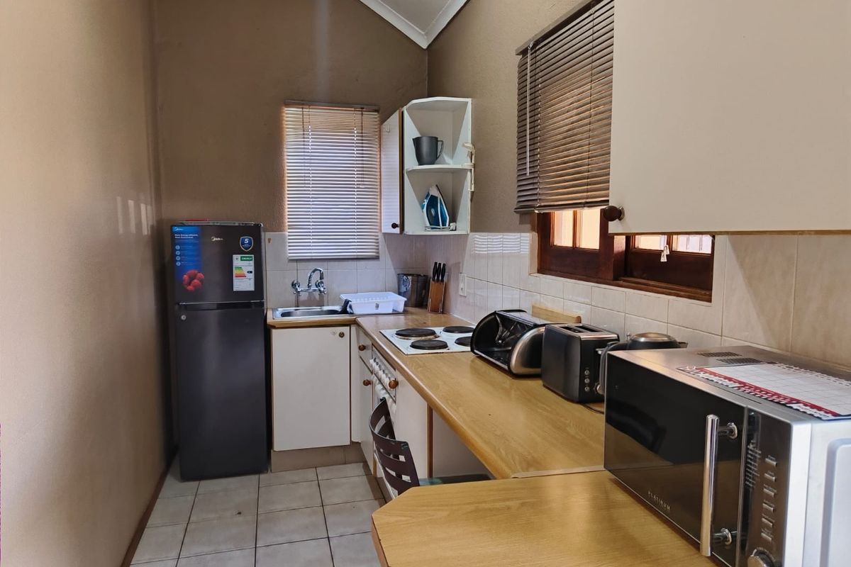 A compact kitchen featuring laminate countertops, built-in cabinetry, an electric hob, and a stainless steel sink with tiled backsplash and venetian blinds.