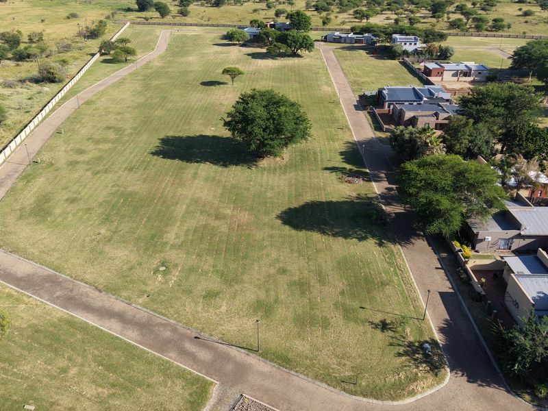 Aerial view of a large, well-maintained vacant residential plot featuring established trees, perimeter fencing, and paved access roads within a residential estate.