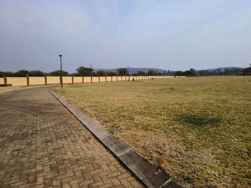 A spacious vacant residential plot featuring a paved driveway, perimeter wall with electric fencing, and established grass area.