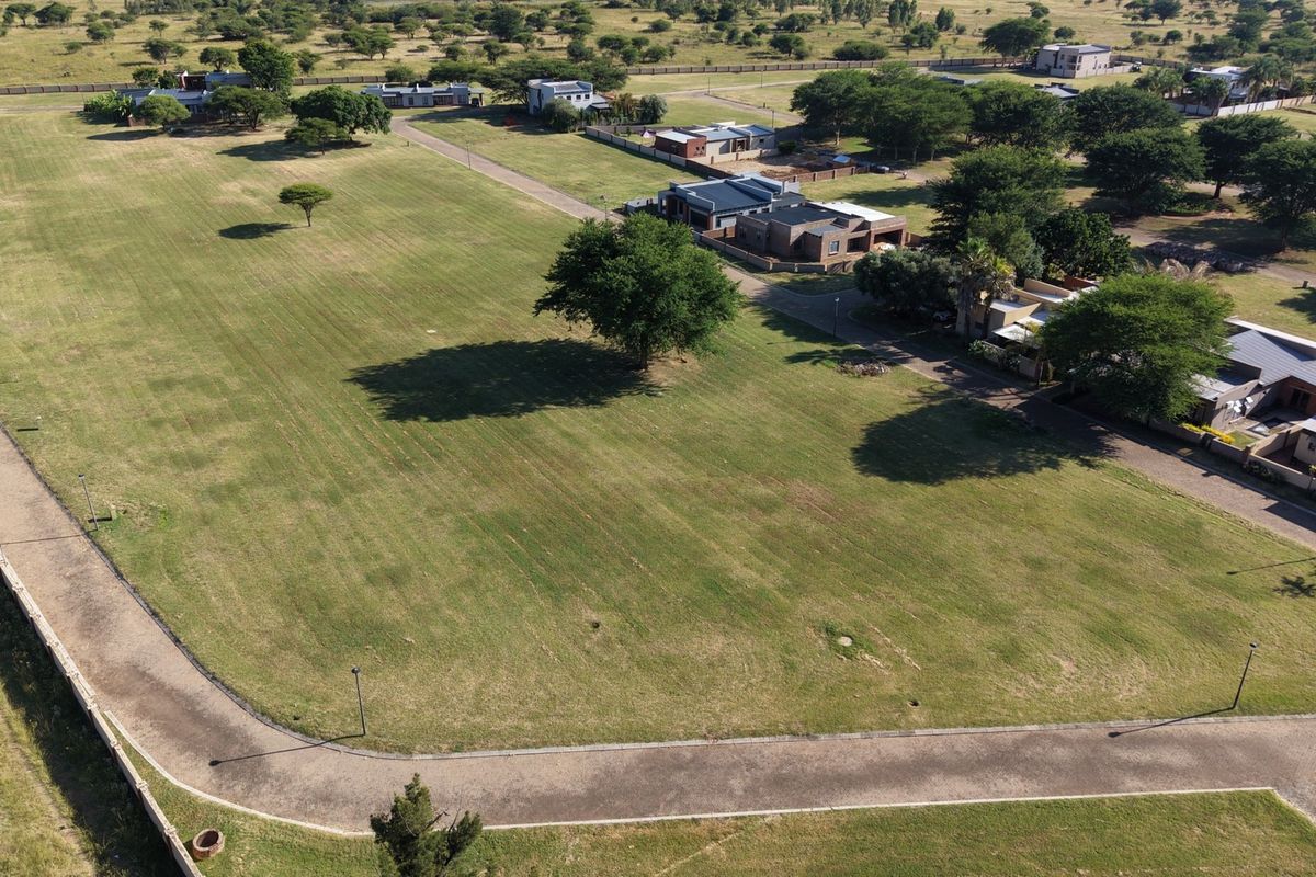 An aerial view of a vacant residential plot featuring well-maintained grass, surrounding infrastructure, and established trees within a residential estate.
