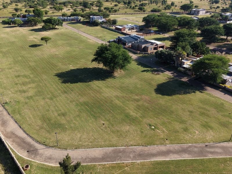 An aerial view of a vacant residential plot featuring well-maintained grass, surrounding infrastructure, and established trees within a residential estate.