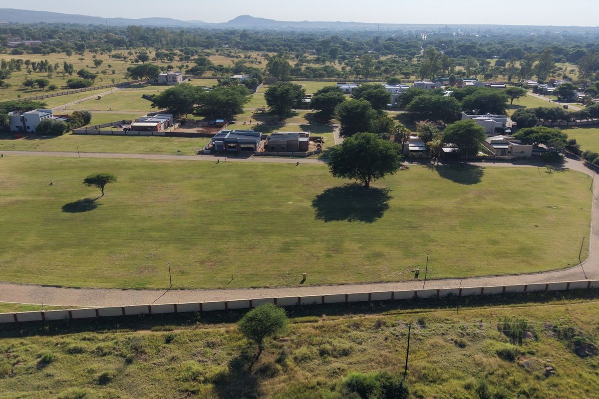 An aerial view of a large, level residential vacant land plot featuring established grass, perimeter fencing, and paved road access within a developed estate.