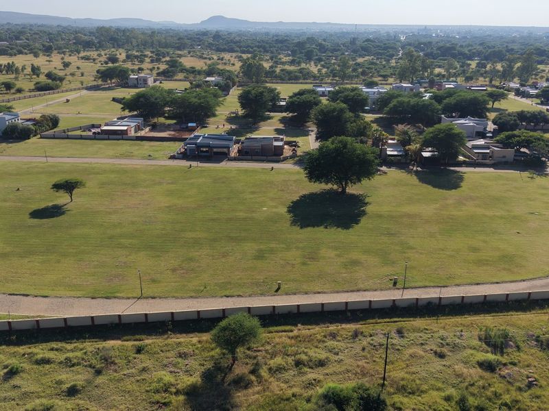 An aerial view of a large, level residential vacant land plot featuring established grass, perimeter fencing, and paved road access within a developed estate.