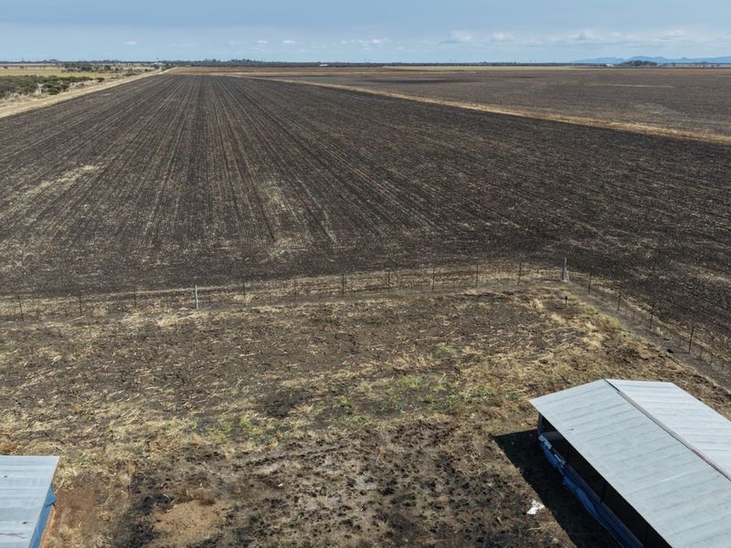 Aerial view of a large, flat agricultural farm property featuring ploughed fields, wire fencing, and two outbuildings with corrugated metal roofs.