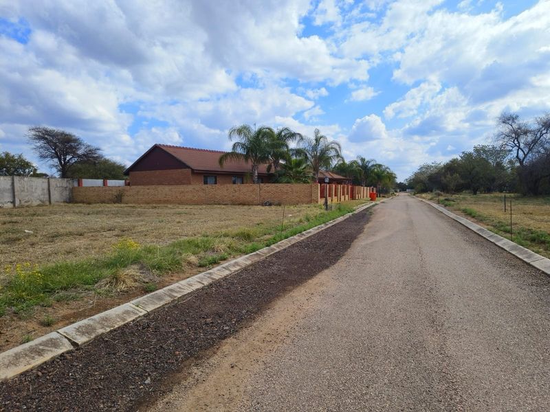 A street view showing a paved road with concrete curbing, adjacent to a vacant residential plot and a brick-walled property.