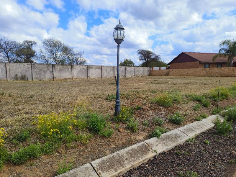 A vacant residential plot featuring a perimeter wall, a central lamp post, and a concrete curb bordering the street.