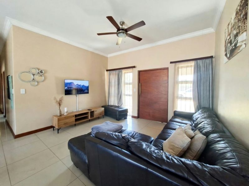 A well-maintained living room featuring tiled flooring, a ceiling fan, and a solid wood entrance door with natural light from large windows.
