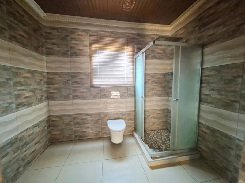 A spacious bathroom featuring full-height decorative wall tiling, a glass shower enclosure, a wall-mounted toilet, and a timber-paneled ceiling.