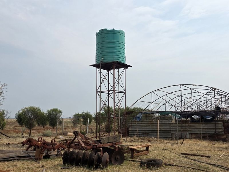Farm exterior featuring a raised water tank on a steel stand and a dilapidated metal-framed structure with corrugated cladding.