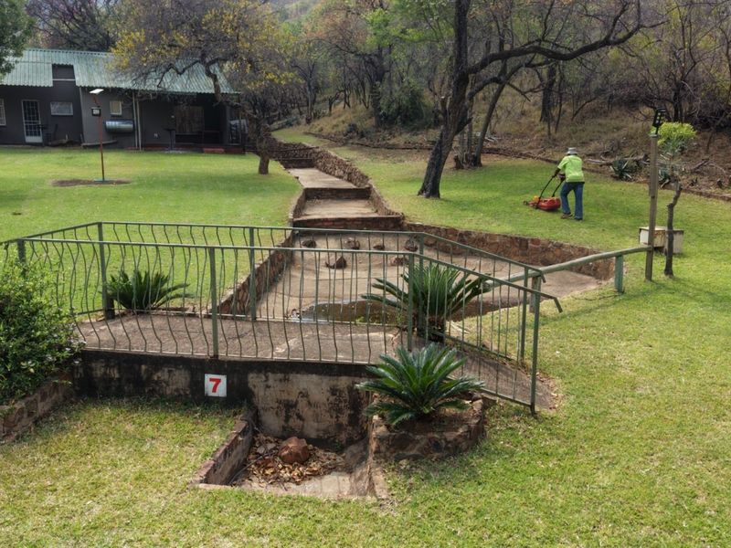 A spacious farm garden featuring a metal bridge over a stone-lined drainage channel, surrounded by mature trees and well-maintained lawn areas.