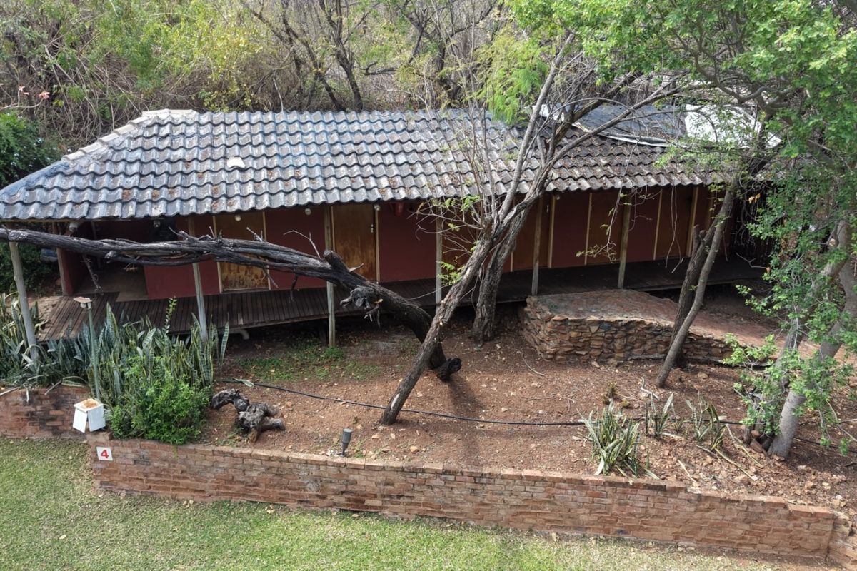 An elevated view of a rustic farm-style dwelling featuring a tiled roof, wooden deck, and brick retaining wall set within a natural bushveld environment.