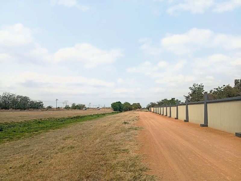 A view of a vacant residential plot featuring a dirt access road and a long perimeter boundary wall.
