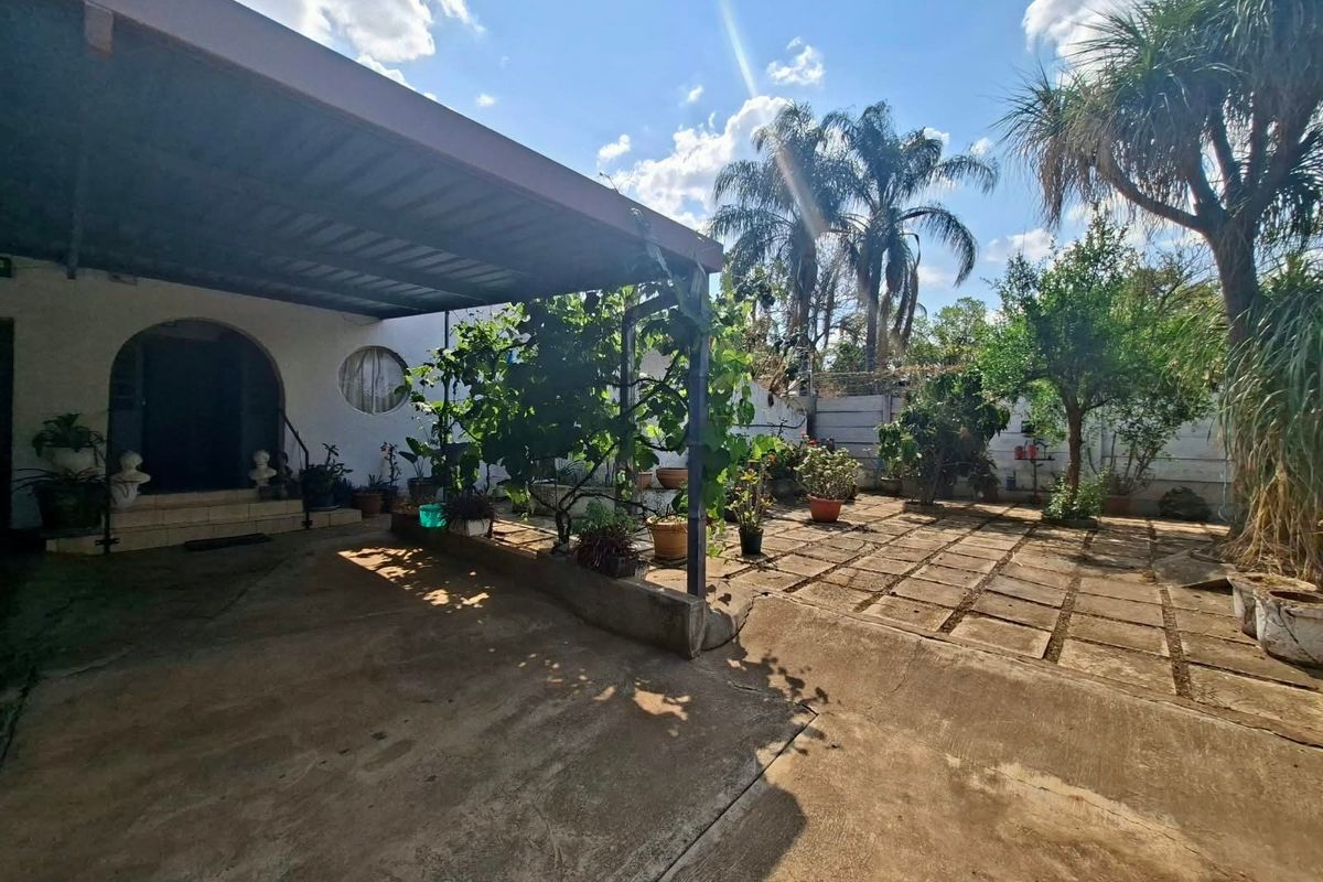 A rear exterior view featuring a covered patio area with concrete paving, an arched entrance, and a landscaped garden with mature trees.