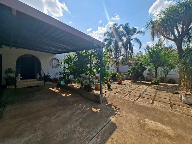 A rear exterior view featuring a covered patio area with concrete paving, an arched entrance, and a landscaped garden with mature trees.