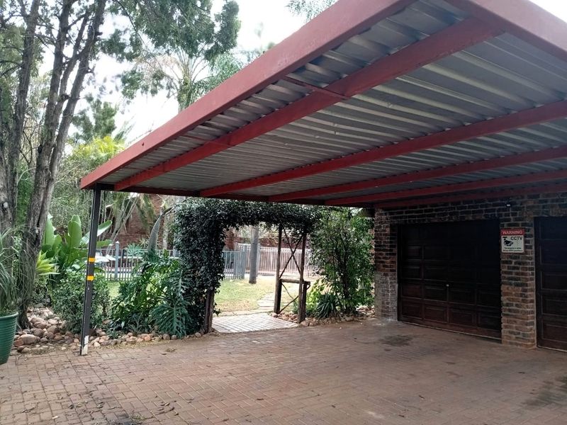 A covered carport area with a paved driveway and brick garage structure, featuring a garden entrance and mature landscaping.