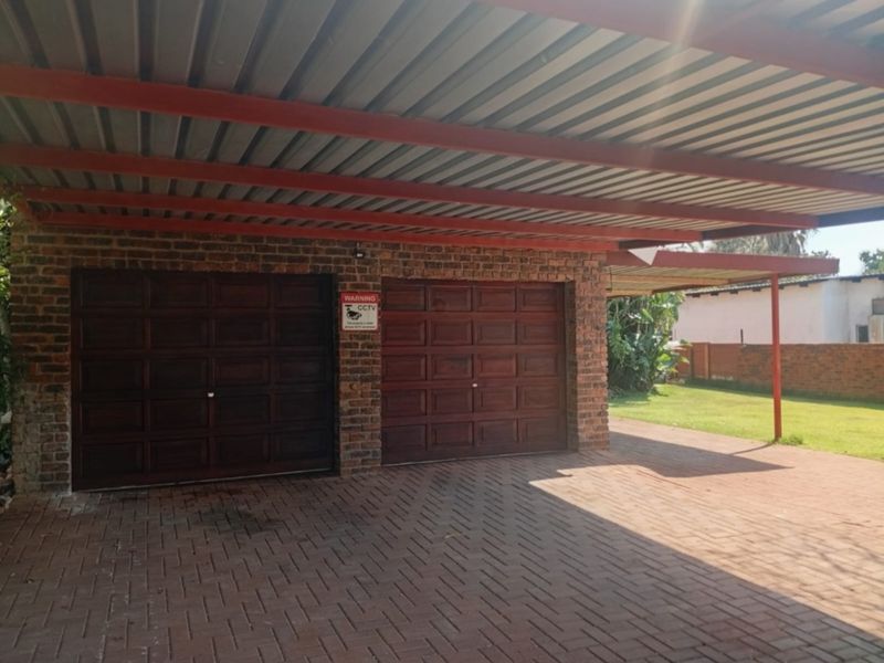 A double garage with wooden doors and an attached carport, featuring a brick exterior and paved driveway area.